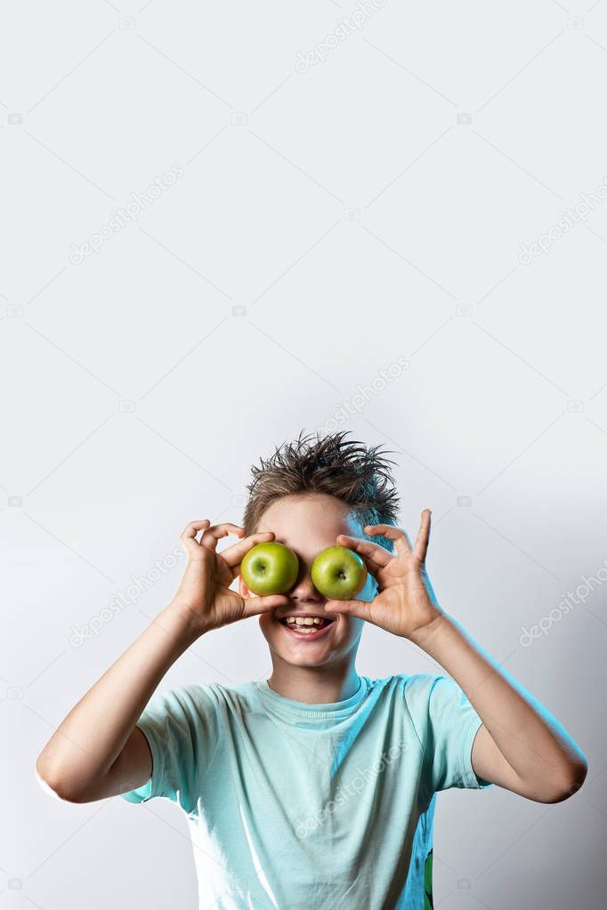 boy in a blue t-shirt put two green apples to his eyes and laughs on a light background