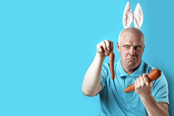 Hombre calvo y brutal con camiseta ligera y orejas de conejo. En sus manos sostiene la zanahoria de un tamaño diferente . — Foto de Stock