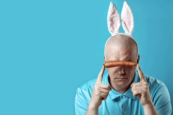 Un hombre calvo y brutal con una camisa ligera y orejas de conejito. En las manos de él sostiene una zanahoria, que cierra los ojos — Foto de Stock