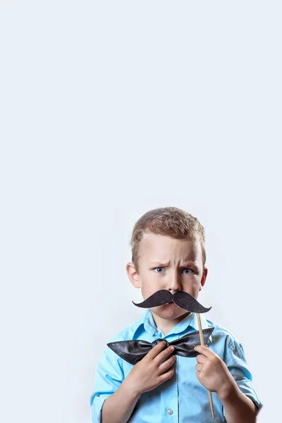 Franzindo a testa menino sério em uma camisa leve colocar um bigode em um pau e um laço gravata em seu rosto para fazê-lo parecer mais velho . — Fotografia de Stock