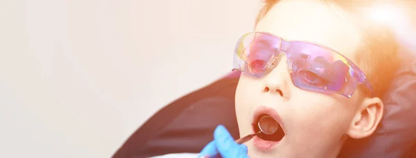 Um rapaz com óculos na cadeira dentária. O médico examina a cavidade oral com um espelho dentário especial — Fotografia de Stock