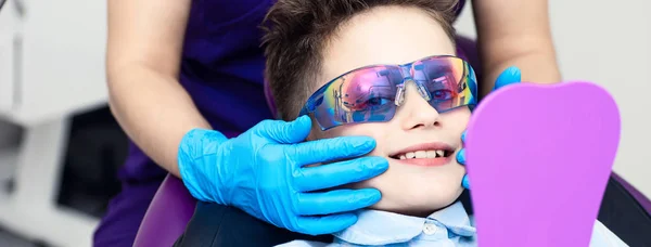 En dreng i særlige briller i tandlægens stol. lægen i sterile handsker viser ham sit smil i spejlet - Stock-foto