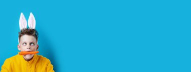 şaşılık ve tavşan kulakları ile sarı bir ceket çocuk, mavi bir arka plan üzerinde ağzında bir havuç tutarak