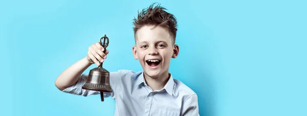 Um menino feliz de camisa leve vai para a escola. Ele tem um sino na mão, que toca e sorri . — Fotografia de Stock