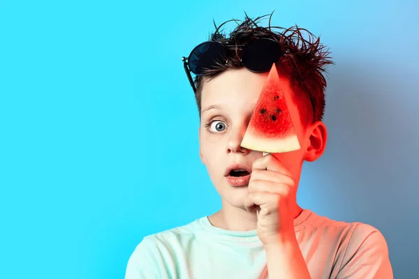Dreng i lys t-shirt vandmelon på en pind lukker det ene øje på en blå baggrund - Stock-foto