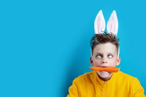 Хлопчик у жовтій куртці зі стрічкою та вухами заєць, тримає моркву в роті на синьому фоні — стокове фото