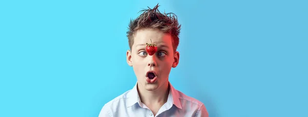 Menino surpreso com um morango caindo guinou seus olhos para o nariz em um fundo azul . — Fotografia de Stock