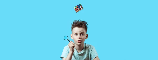 Un niño con una camiseta ligera sostiene una pequeña lupa en su mano y un cubo de Rubik cae desde arriba . — Foto de Stock