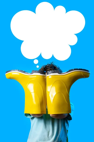 A boy in a light t-shirt covered his face with yellow rubber boots. symbolizes autumn on blue background — Stock Photo, Image