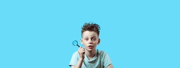 Um menino em uma camiseta leve segurando uma pequena lupa no fundo azul — Fotografia de Stock