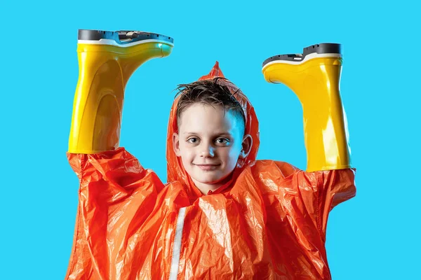 Усміхнений хлопчик в помаранчевому плащі застряг у гумових чоботях на синьому фоні — стокове фото