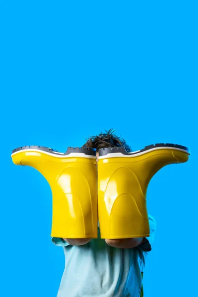 Un niño con una camiseta ligera se cubrió la cara con botas de goma amarillas. simboliza el otoño sobre fondo azul — Foto de Stock