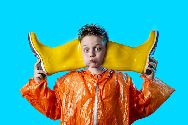Sjov dreng i en orange frakke med pustede kinder og gummistøvler på blå baggrund - Stock-foto