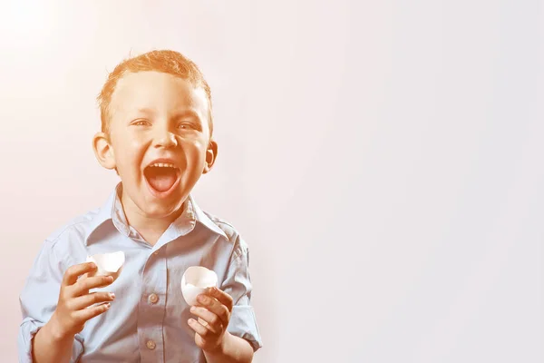 Dreng i en lys skjorte holder en skal fra ægget og smiler på en lys baggrund - Stock-foto