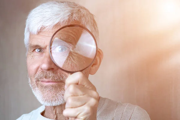 A gray-haired man in light clothes looking through a magnifying glass — Stock Photo, Image