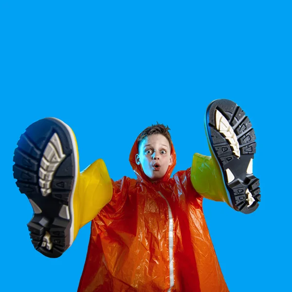 Усміхнений хлопчик в помаранчевому плащі застряг у гумових чоботях на синьому фоні — стокове фото