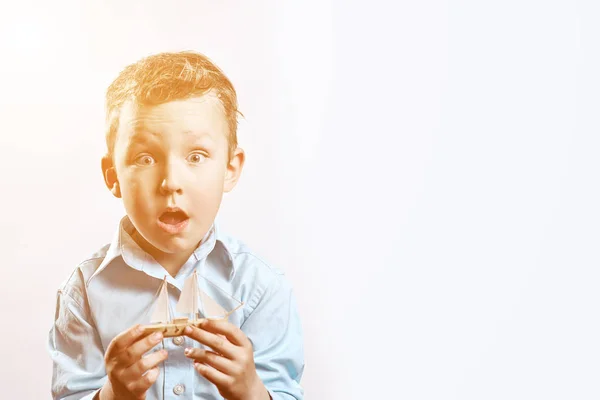 A surprised boy in a blue shirt holding a boat in his hands on a light background — Stock Photo, Image