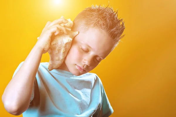 Un niño en una camiseta ligera escucha el mar en una concha de mar sobre un fondo de color — Foto de Stock