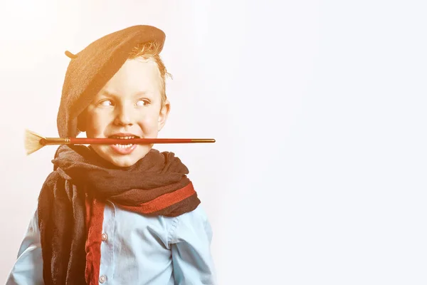 Junge Künstler in schwarzer Baskenmütze, Schal und mit Pinsel im Mund auf hellem Hintergrund — Stockfoto