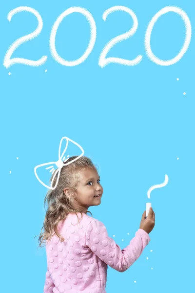 Cute girl with white chalk on a blue background. Writes on the Board 2020 year — Stock Photo, Image