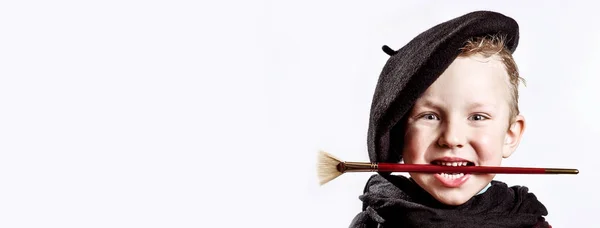 Niño artista en boina negra, bufanda y con un cepillo en la boca sobre un fondo claro — Foto de Stock