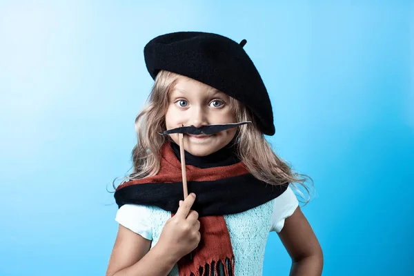 Chica divertida en boina negra, bufanda y bigote en un palo sobre fondo azul — Foto de Stock