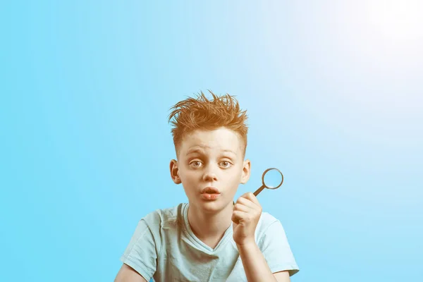 Um menino em uma camiseta leve segurando uma pequena lupa no fundo azul — Fotografia de Stock