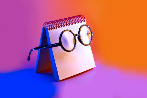 Fekete kerek szemüveg feküdjön egy jegyzettömb a Neonfény a rózsaszín háttér — Stock Fotó