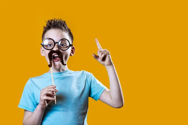 Um menino em uma camiseta leve com bigode e óculos segura seu dedo indicador contra fundo brilhante — Fotografia de Stock