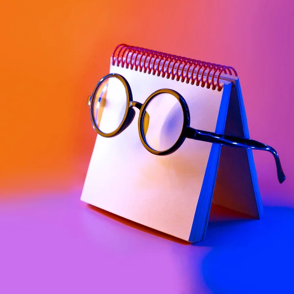 Fekete kerek szemüveg feküdjön egy jegyzettömb a Neonfény a rózsaszín háttér — Stock Fotó