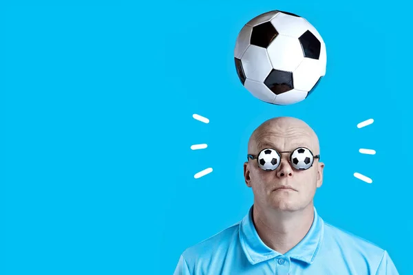 Calvo hombre brutal en gafas redondas oscuras con reflejos. Un balón de fútbol sobrevolaba. Sobre un fondo azul . — Foto de Stock