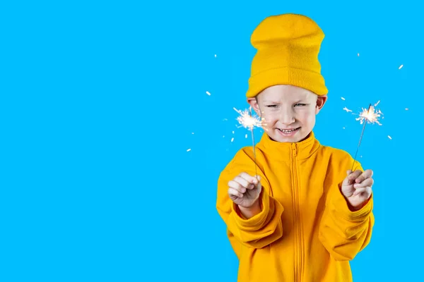 Ett litet glad barn i en gul hatt och jacka håller brinnande tomtebloss i händerna på blå bakgrund — Stockfoto