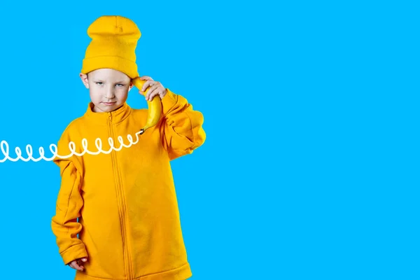 Parlak sarı ceketli ve şapkalı havalı çocuk elinde telefon gibi bir muz tutuyor. Mavi arkaplanda — Stok fotoğraf