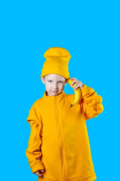 Chico fresco con una chaqueta de color amarillo brillante y sombrero sostiene un plátano en sus manos como un teléfono. Sobre fondo azul — Foto de Stock