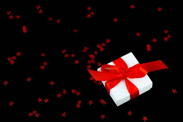Confezione regalo bianca con un nastro rosso e una dispersione di stelle su sfondo nero . — Foto Stock