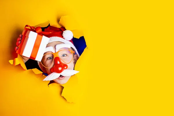 Little cheerful Santa in glasses with red nose and mustache gives a gift, getting out of the ragged yellow background — Stock Photo, Image