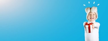 Beyaz tavşan kostümlü neşeli bir çocuk mavi arka planda kırmızı kurdeleli bir hediye veriyor.