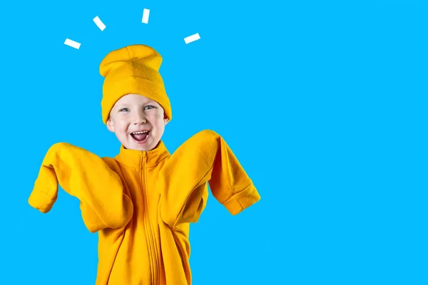 Un chico alegre con una chaqueta de color amarillo brillante y sombrero se regocija con entusiasmo sobre fondo azul — Foto de Stock