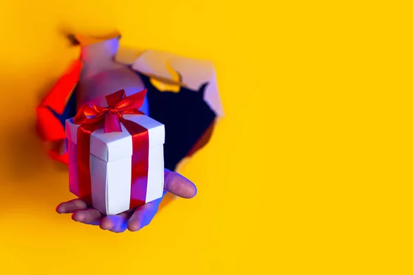 Um presente com um arco vermelho na mão emerge de um furo irregular no fundo de papel amarelo, iluminado por luz de néon — Fotografia de Stock