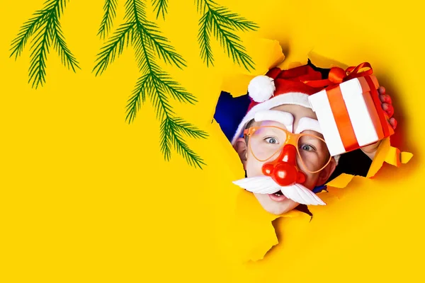 Mały wesoły Mikołaj w okularach z czerwonym nosem i wąsami daje prezent, wychodząc z poszarpanego żółtego tła — Zdjęcie stockowe
