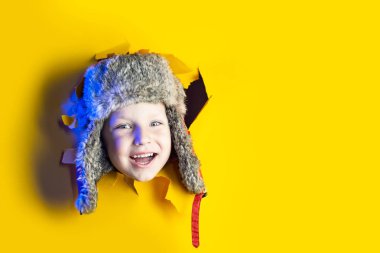 Sarı karton arka plandaki bir delikten kulaklık takan, kışlık şapkalı küçük neşeli bir çocuk.