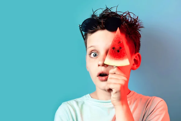 Dreng i lys t-shirt vandmelon på en pind lukker det ene øje på en blå baggrund - Stock-foto