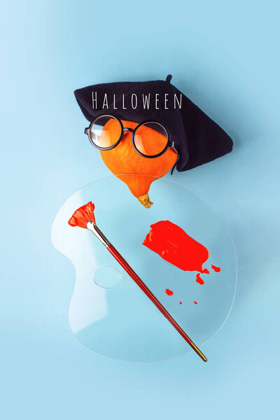 Orange decorative pumpkin artist in black beret, glasses, with a brush, palette and red paint on a blue background. Halloween concept.