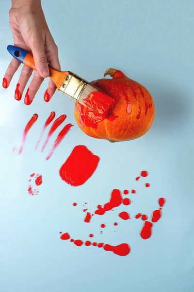 De hand met de borstel verft de pompoen rood. Verf is als bloed. Nabijgelegen plekjes, druppels, sprays, vlekken, spatten. Halloween concept. — Stockfoto