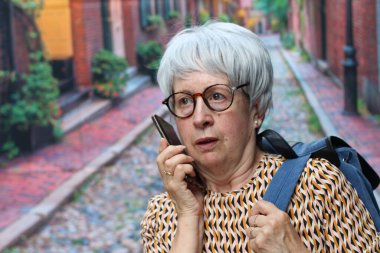 Sokakta telefonla konuşan gri saçlı, sırt çantalı olgun bir kadının portresi.