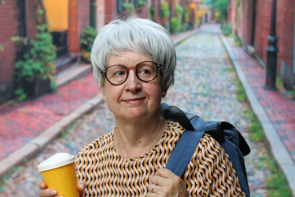 portrait of mature woman with grey hair in casual clothes with backpack and paper cup of coffee on street