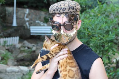 Steampunk gözlükleri ve altın maskeli kedi tutan yakışıklı genç adamın yakın plan portresi.