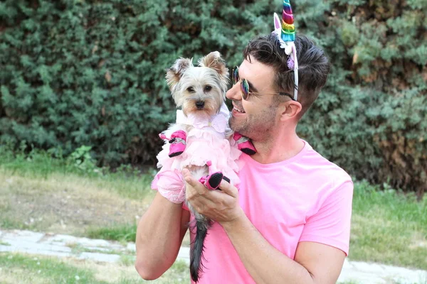 身穿粉色T恤的年轻帅气男子与他的小狗在花园里的独角兽角的特写 — 图库照片