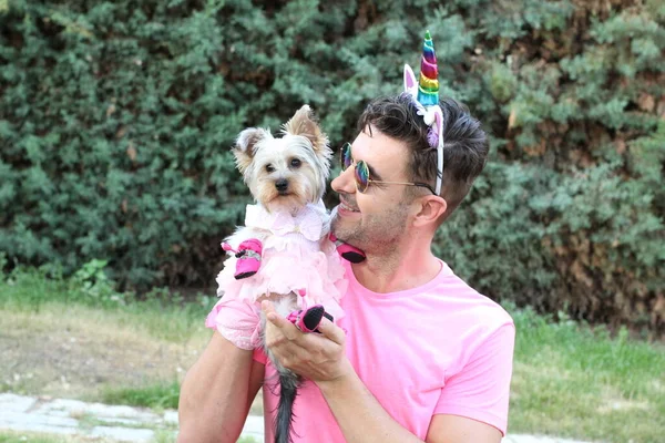 身穿粉色T恤的年轻帅气男子与他的小狗在花园里的独角兽角的特写 — 图库照片