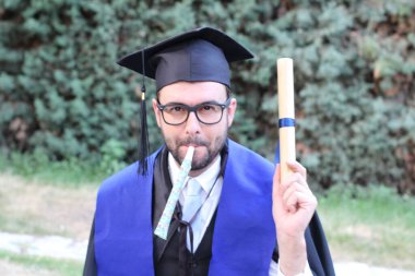 Mezuniyet kıyafetleri içinde, sarılmış diploması ve parkta piposu olan yakışıklı bir gencin yakın plan portresi.